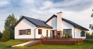A rear exterior view of a modern, ranch-style home showcasing key tiny home features: a large elevated wooden deck, an outdoor fireplace, and expansive glass sliding doors.