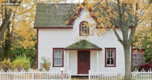 Charming classic white 2 story tiny home with dark red window trim, a small front porch, and a white picket fence under autumn trees.