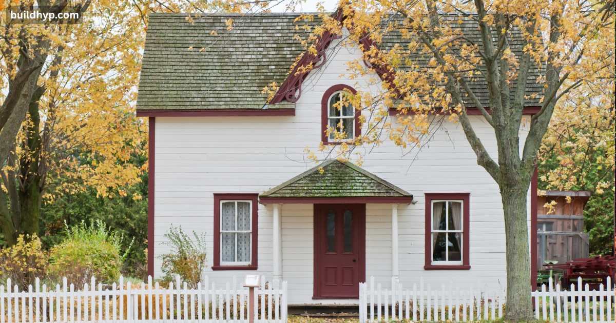 Charming classic white 2 story tiny home with dark red window trim, a small front porch, and a white picket fence under autumn trees.