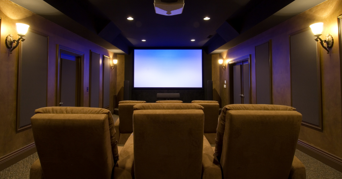 A view from the back of a home theater showing three rows of tan velvet recliners. The room has moody lighting and decorative wall sconces, perfect for home theaters in small cabins.