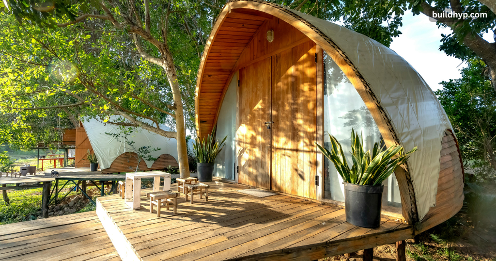 Modern arched small wooden cabin pods set outdoors on a timber deck, surrounded by green trees, potted snake plants, and sunny outdoor seating.