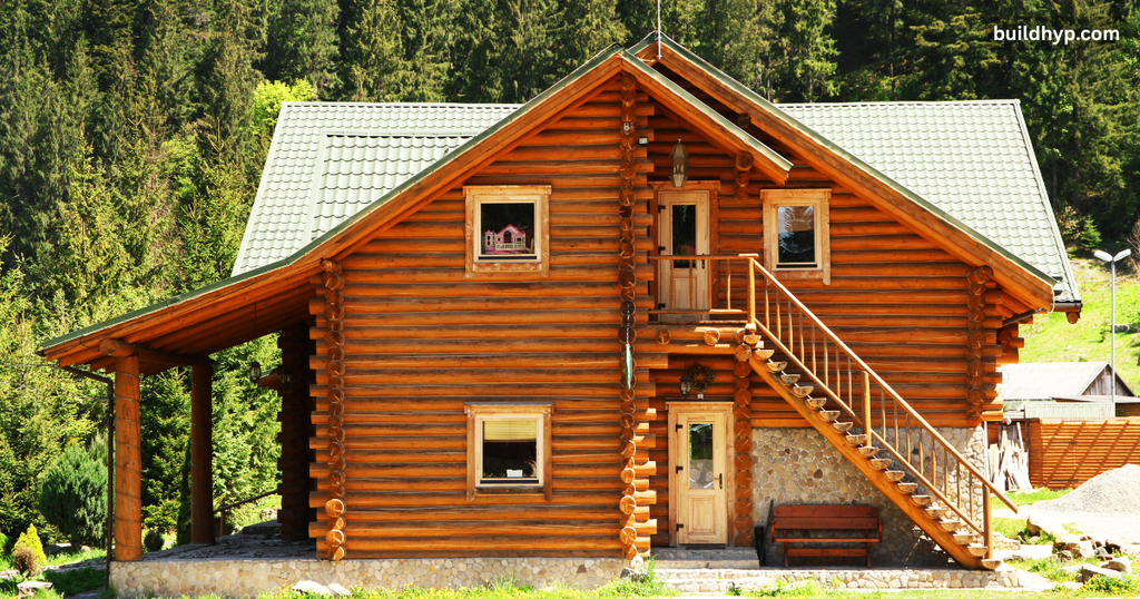 Exterior view of a two-story small wooden cabin built with polished rounded logs, featuring an outdoor wooden staircase, a side porch, and a lush green forest background.