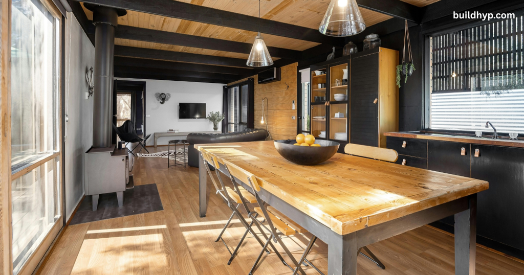Interior of a modern Rocky Mountain tiny house design showing a light wood dining table with black-trimmed chairs, overhead pendant lighting, dark cabinetry, and a small wood-burning stove.