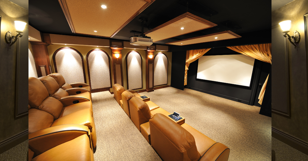 A cozy home theater inside a small cabin, featuring three rows of tan leather recliners, a large projector screen with gold curtains, and warm, recessed ceiling and wall lighting. 