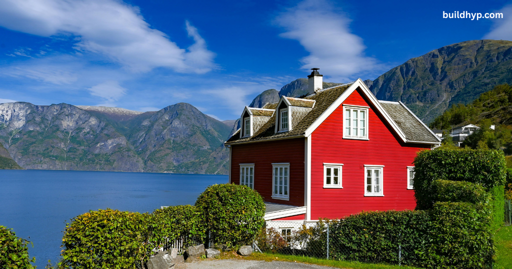 A vibrant red two-story cottage nestled beside a calm blue lake with steep mountain peaks in the background, offering inspiration for scenic Rocky Mountain tiny house builds.