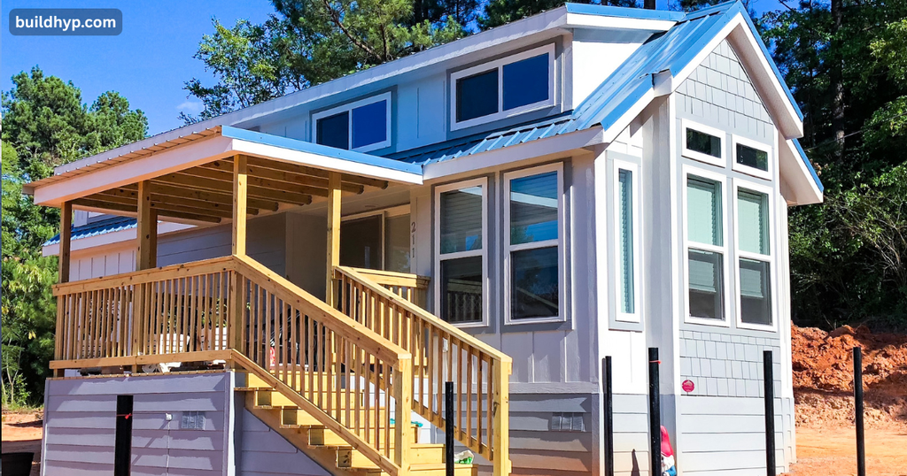 Modern 2 story tiny home on wheels featuring light grey siding, a bright blue metal roof, and an elevated wooden entry porch with stairs.