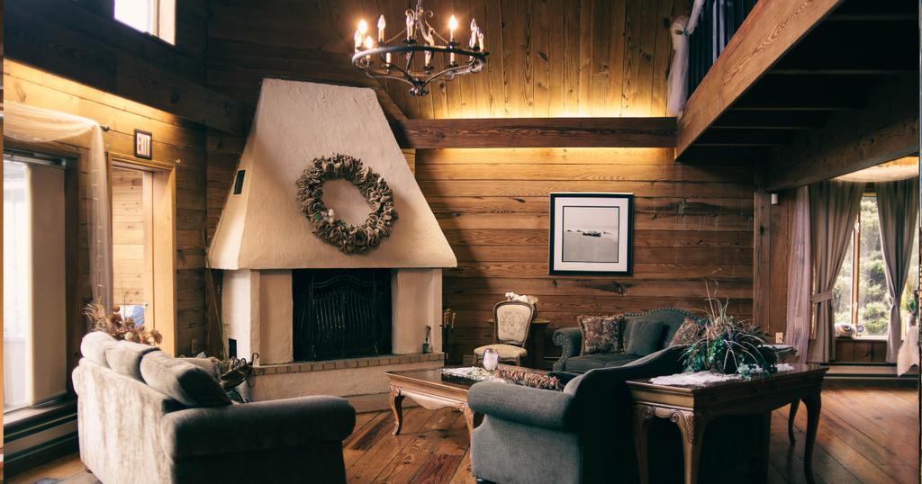 A cozy mountain lodge living room interior with dark wood walls, comfortable armchairs, and a central whitewashed fireplace adorned with a decorative wreath.