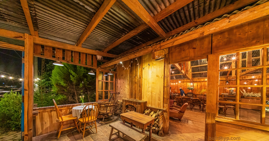 A rustic wood-paneled porch area featuring small dining tables, rocking chairs, and warm string lighting under a corrugated metal roof.