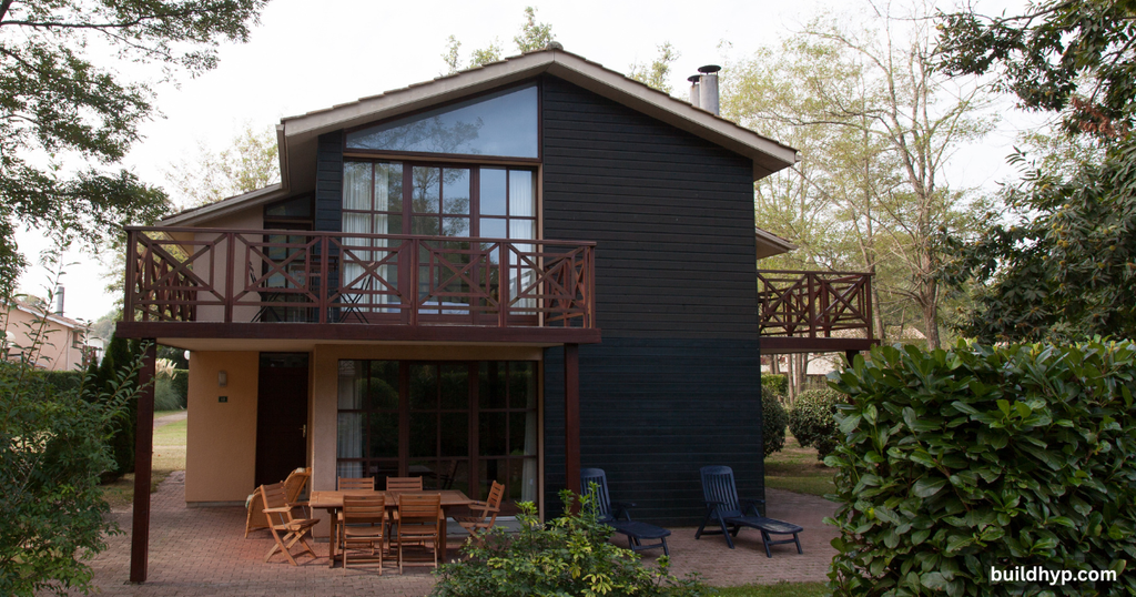 Contemporary dark wood 2 story tiny home featuring a large second-floor balcony, expansive glass windows, and a paved outdoor patio dining area.
