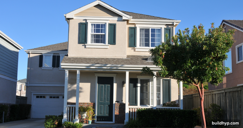 Compact two-story tiny house layout with beige stucco exterior, dark green shutters, a small covered front porch, and a manicured front yard.

