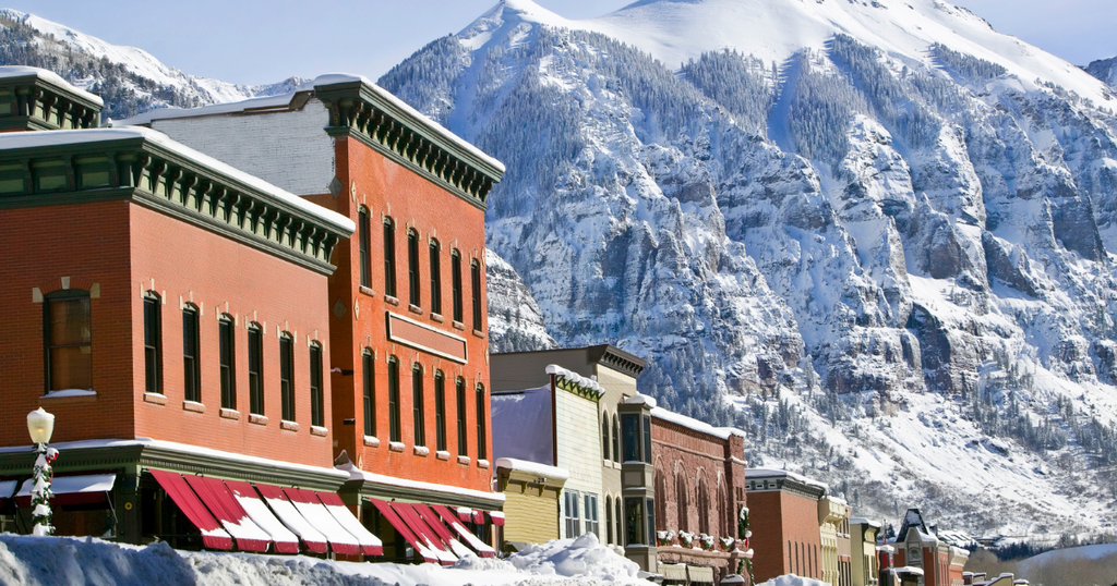 Historic brick buildings line a snow-covered Main Street in Telluride, Colorado, with towering, snow-dusted Rocky Mountains rising dramatically in the background under a clear blue sky.