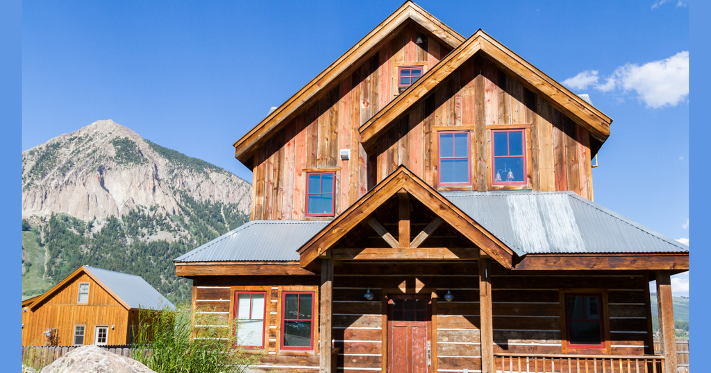 A rustic wooden cabin with red trim windows and a metal roof stands against a backdrop of a majestic, rocky peak, illustrating custom Colorado Mountain Houses built for alpine living.