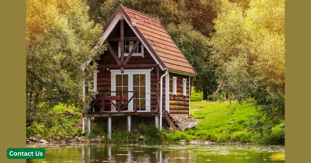 Picturesque small wooden cabin with a red tiled roof elevated on stilts beside a serene pond, surrounded by dense green trees and grass.