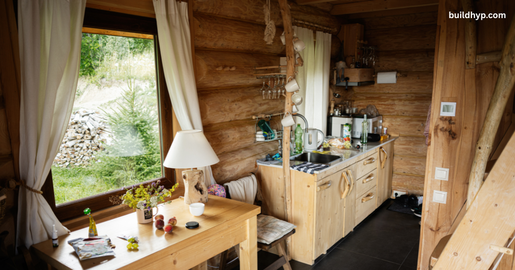 Cozy interior of a rustic small wooden cabin showing a wooden dining table with fresh flowers, a compact kitchen counter, and a large window overlooking pine trees.